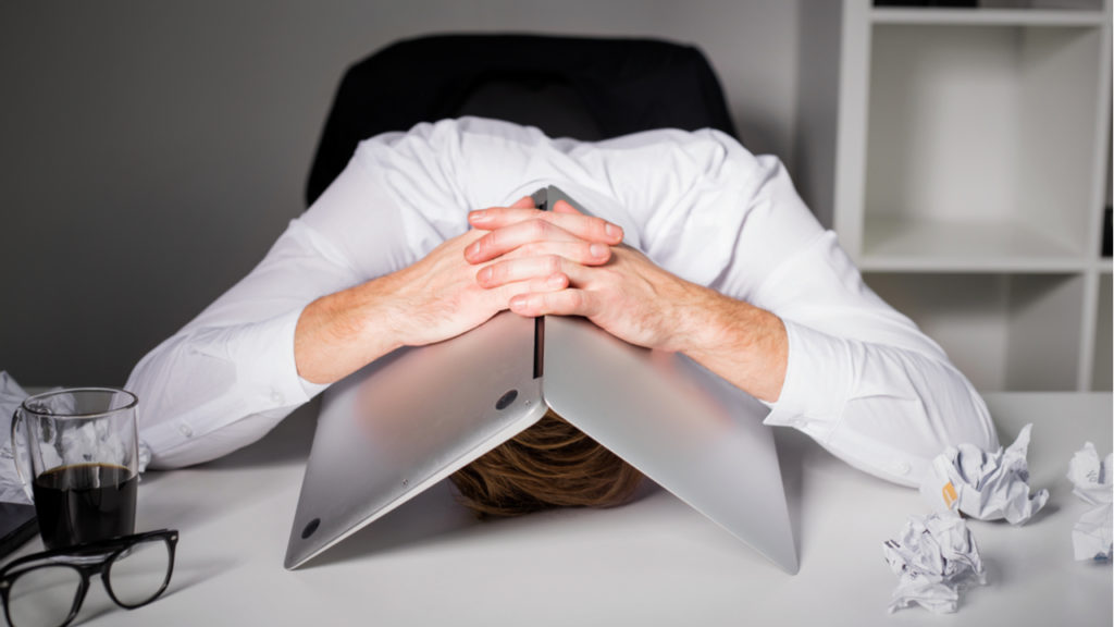 Man face down on desk with lap top over his head in frustration